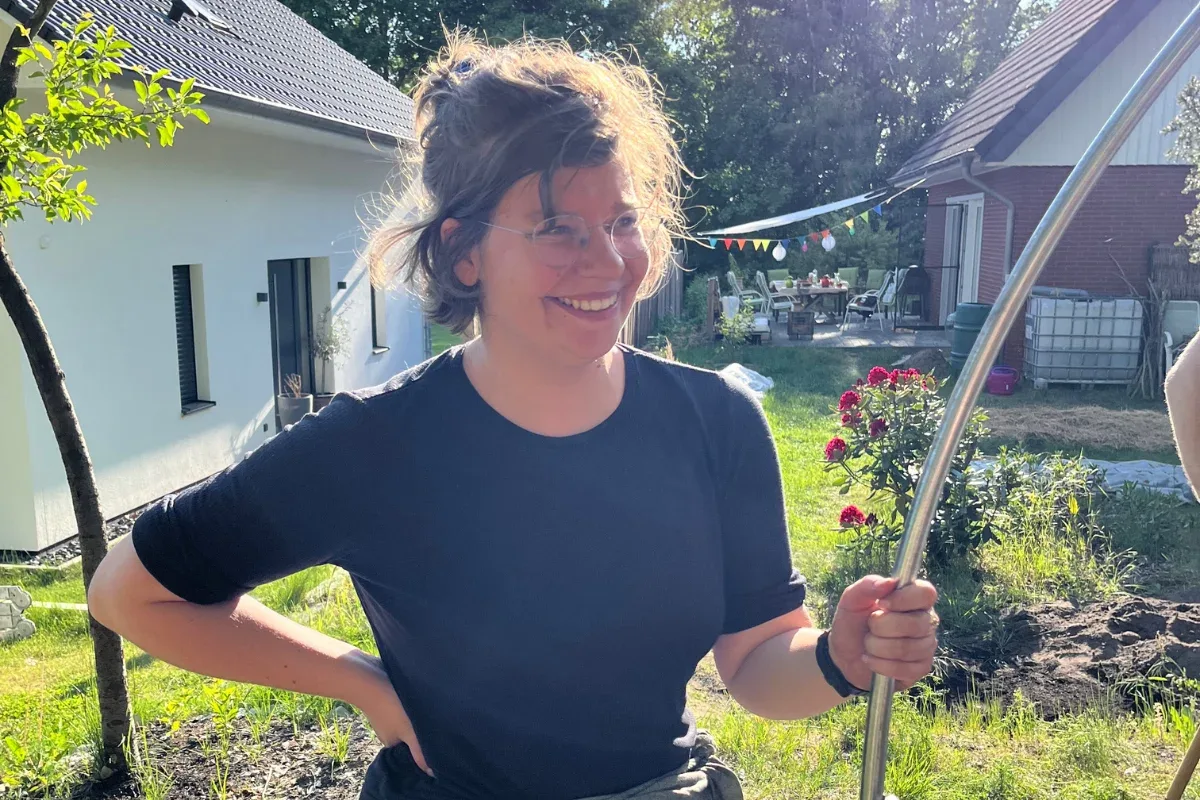 Lachende Frau mit einem Stab in der Hand in einem Garten vor einem Wohnhaus