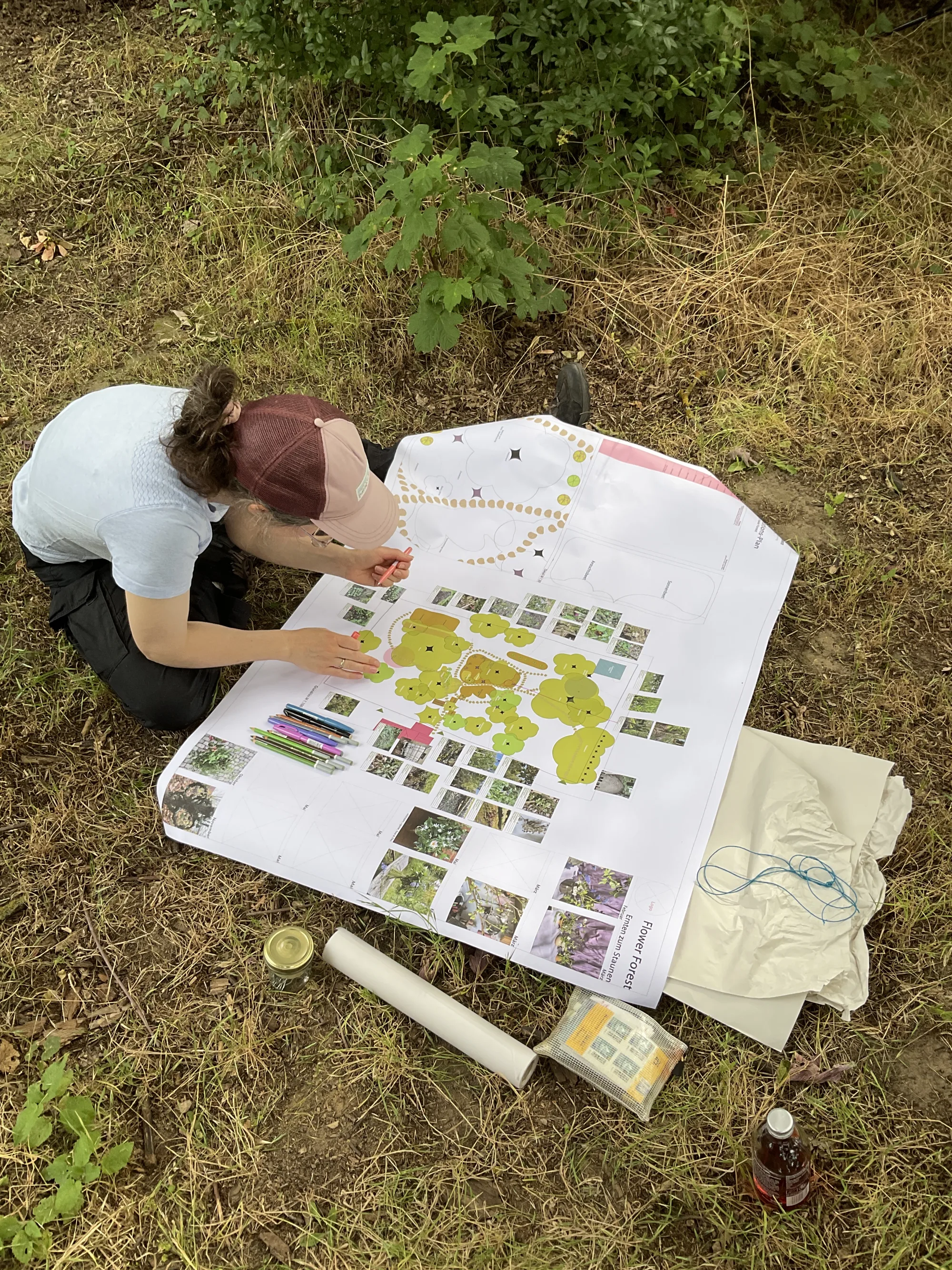 Frau kniet auf der Wiese über einem großen Gartenplan aus Papier mit Pflanzenkarten