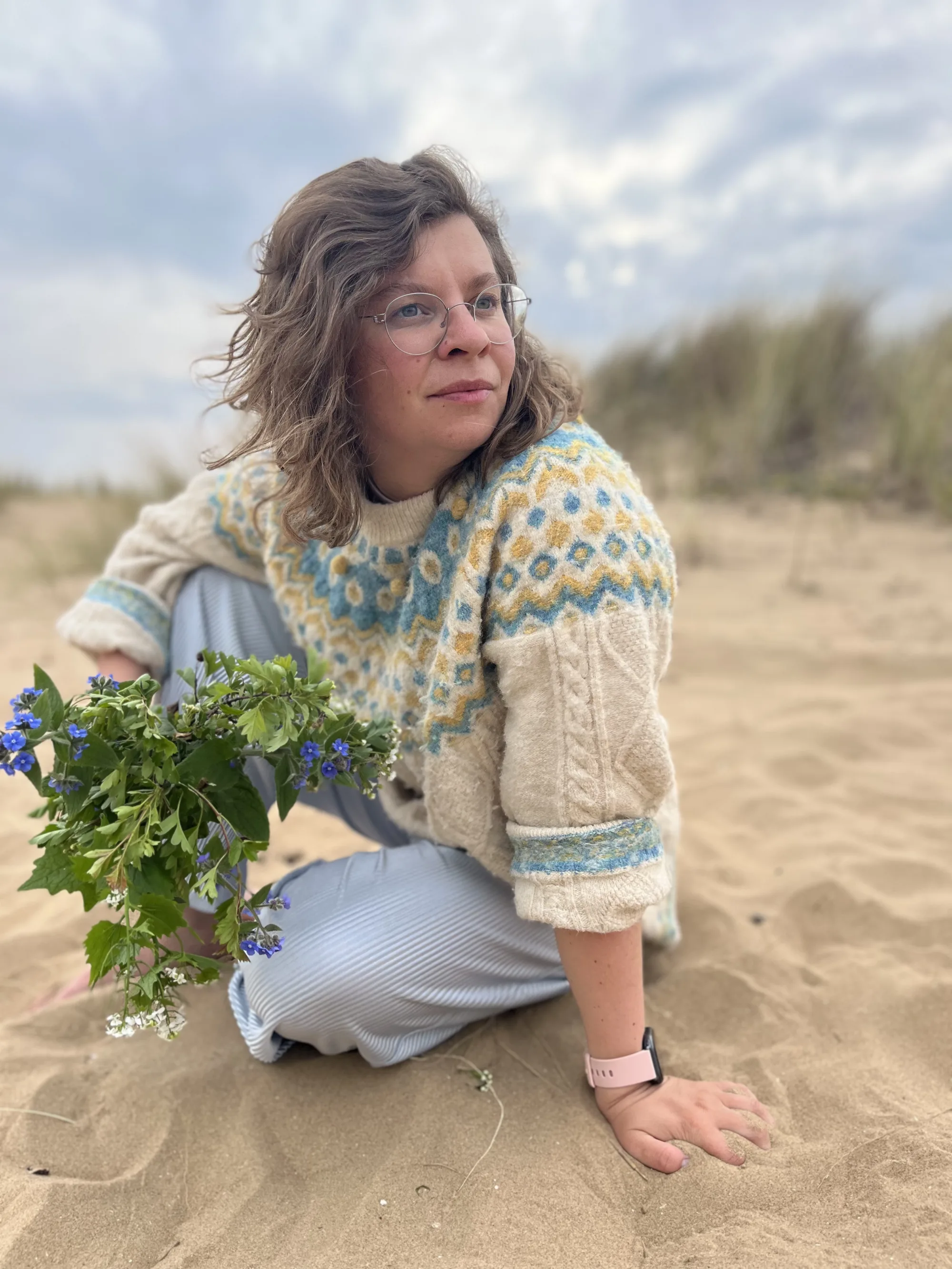 Maureen sitzt im Norwegerpulli im Dünensand, neben sich einen Strauß Wildkräuter, und schaut in die Ferne