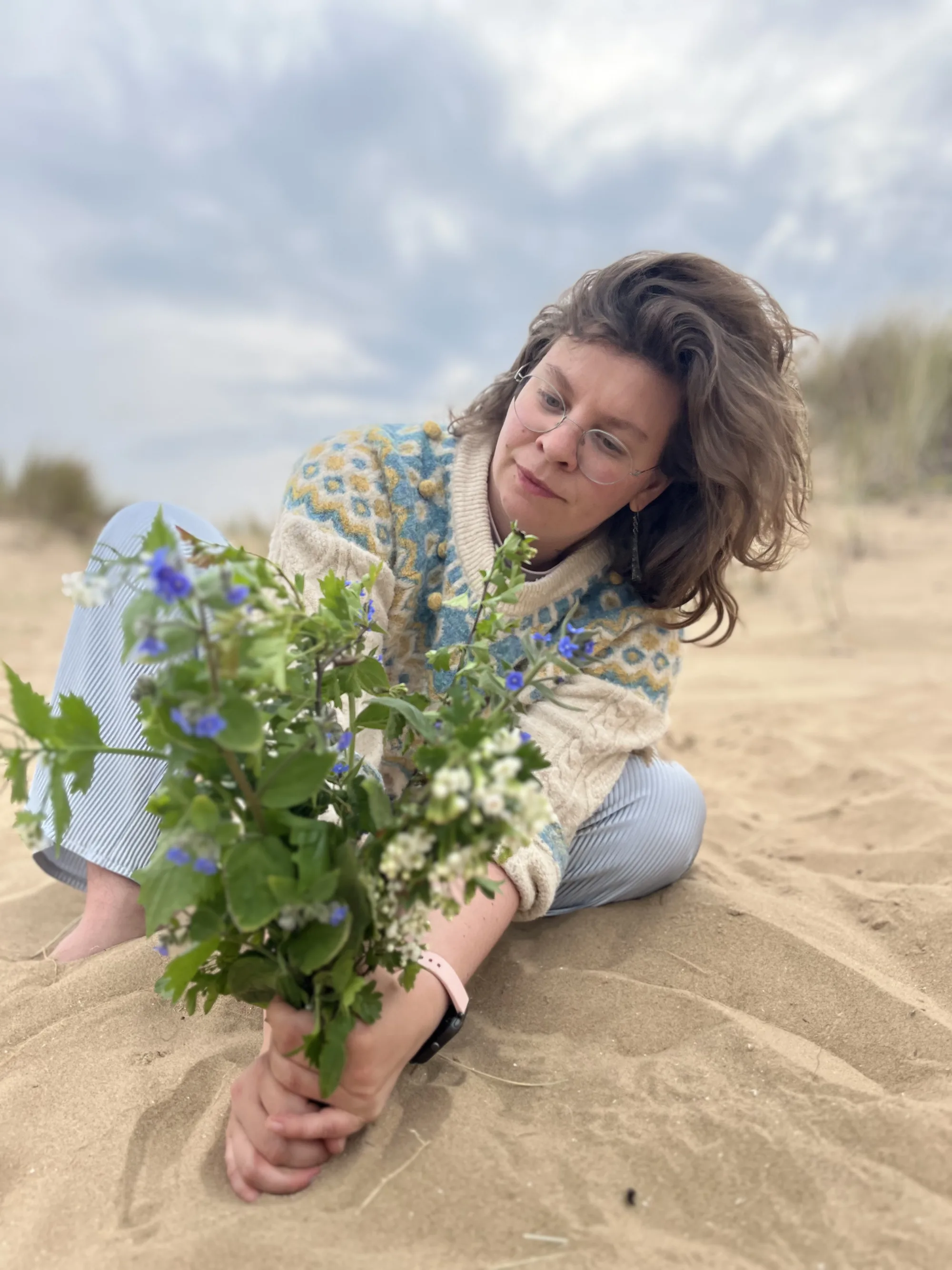 Maureen sitzt mit geschlossenen Augen im Dünensand und hält einen Strauß Wildkräuter