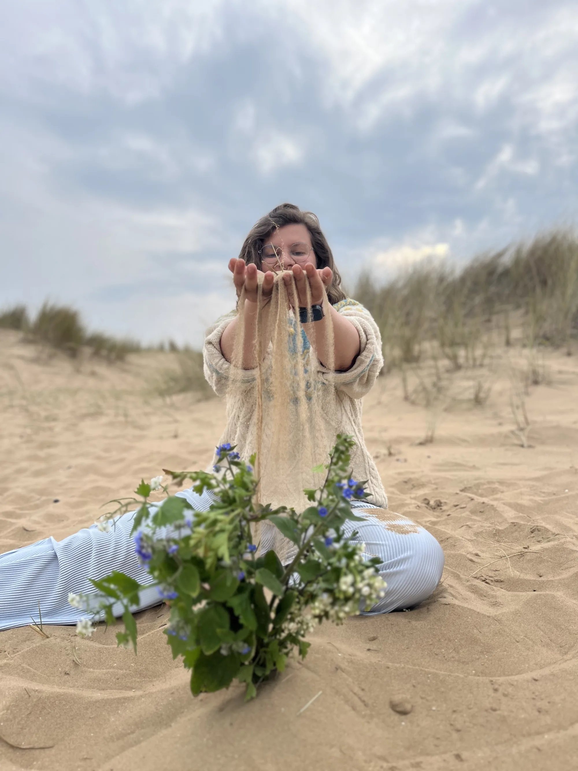 Maureen sitzt im Dünensand und lässt Sand durch ihre Hände rieseln, vor ihr ein Wildkräuterstrauß