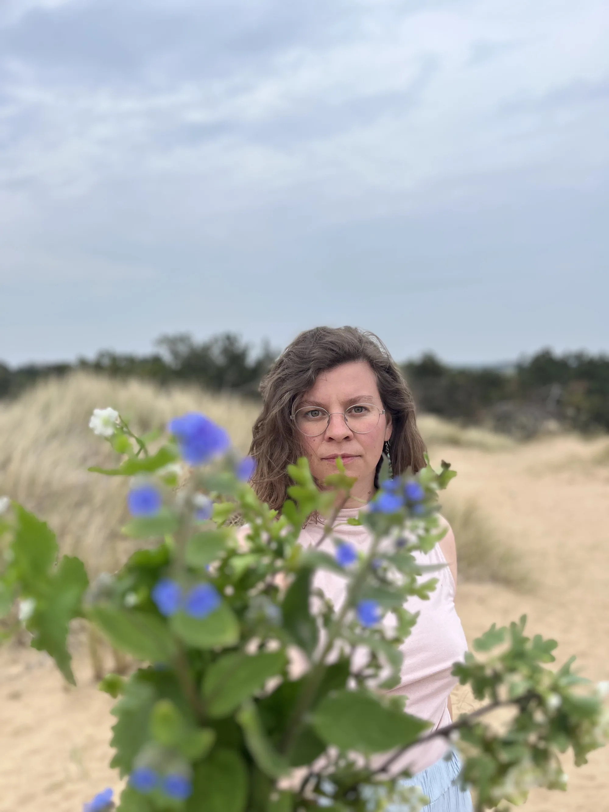 Maureen steht in den Dünen, im Vordergrund unscharf ein Zweig mit blauen Blüten